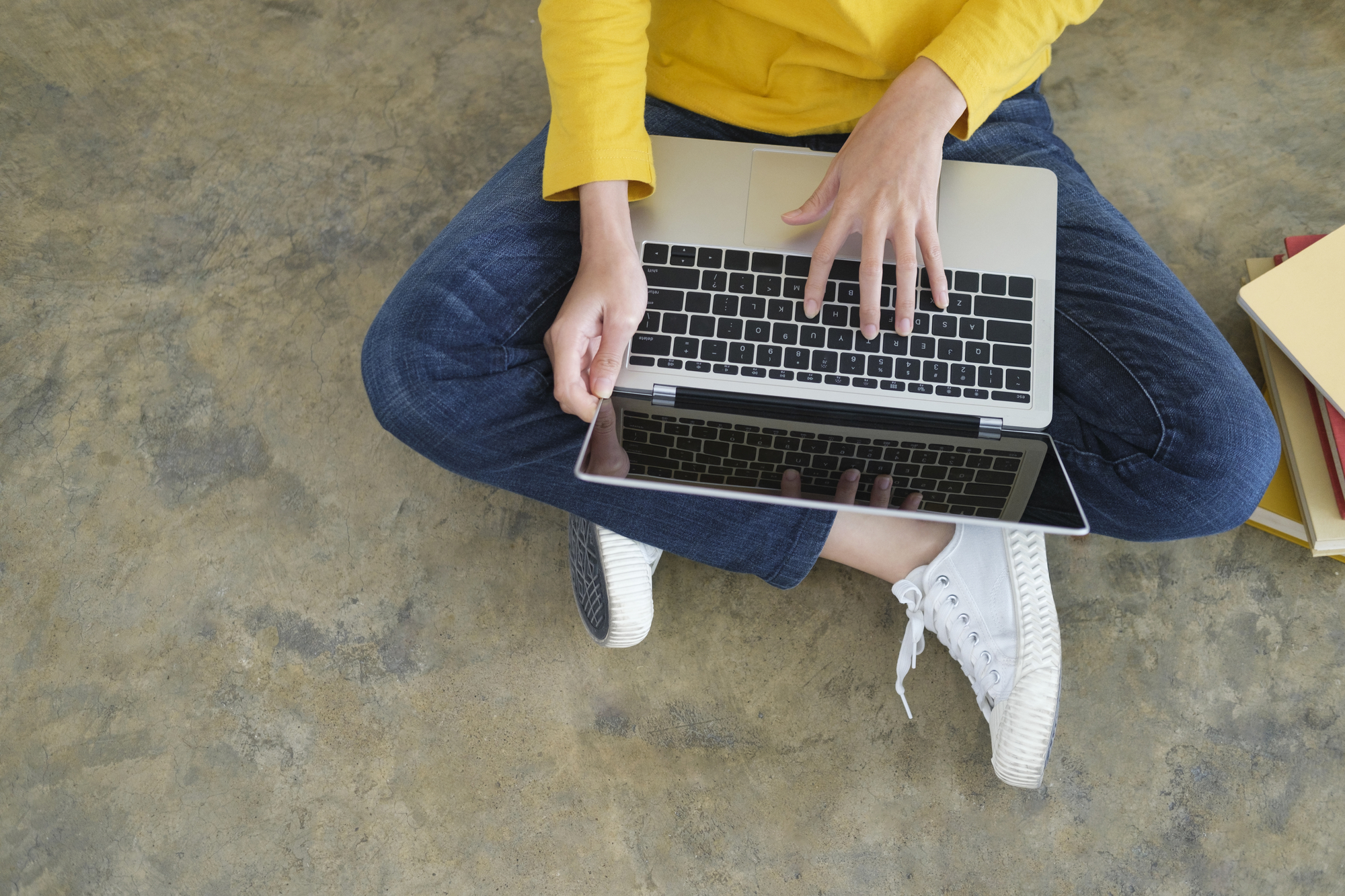 Young woman in yellow top and jeans working or studying online using laptop sitting on the floor.