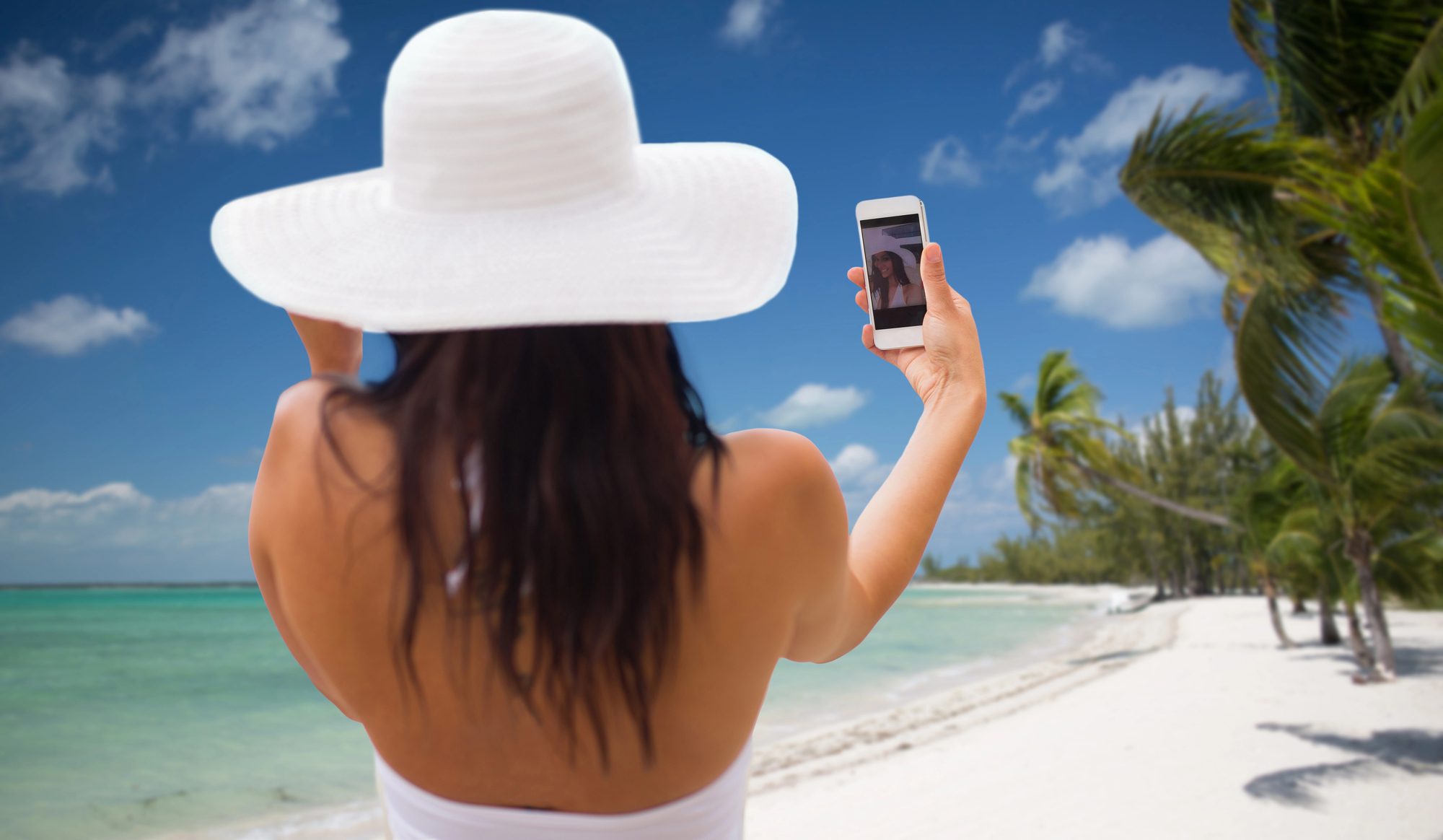 A woman in a white hat takes a selfie on a tropical beach with clear water, white sand, and palm trees under a blue sky, making money taking photos of herself as an influencer