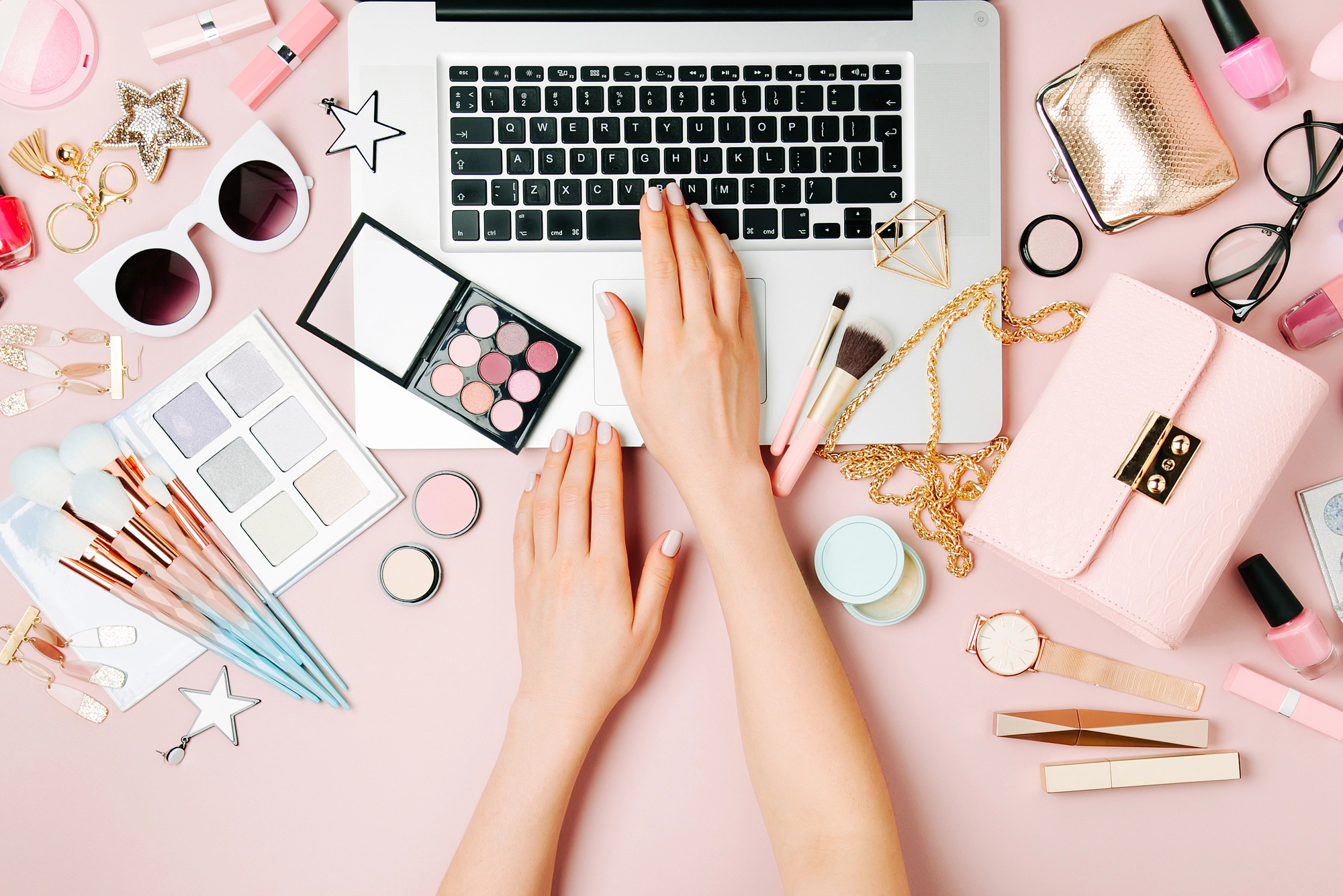 Flat lay image of hands typing on a laptop surrounded by various makeup products and accessories on a pink background, illustrating the vibrant world of influencers and beauty bloggers who get paid to take pictures of themselves.