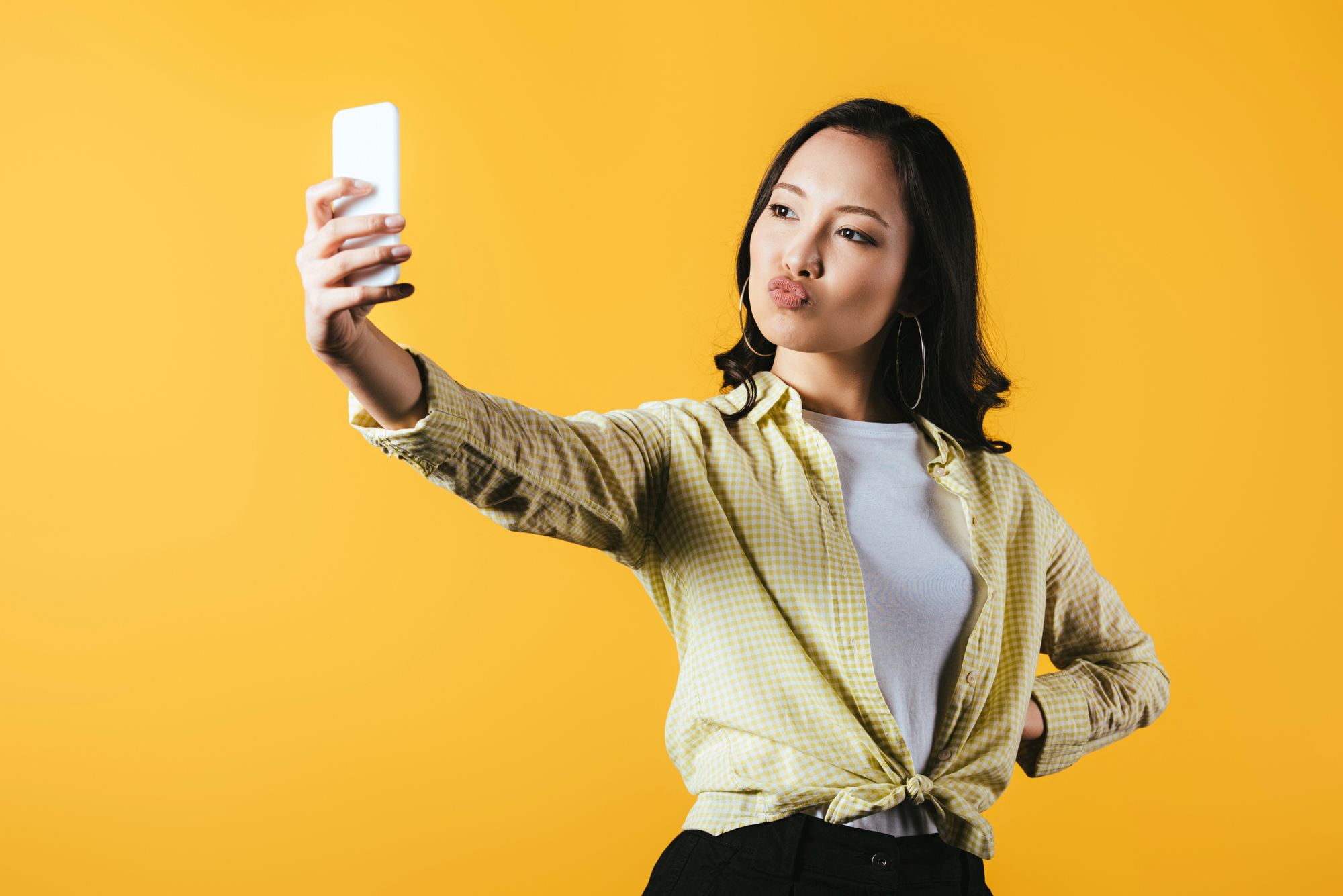 A young woman wearing a yellow checked shirt and hoop earrings takes a selfie against a bright yellow background, showcasing how easy it can be to get paid to take pictures of yourself.