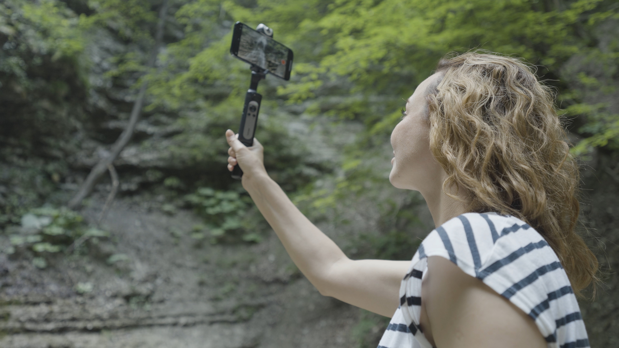 A person with curly hair is taking a selfie in a forest using a smartphone mounted on a handheld stabilizer, embracing the opportunity to make money taking photos.