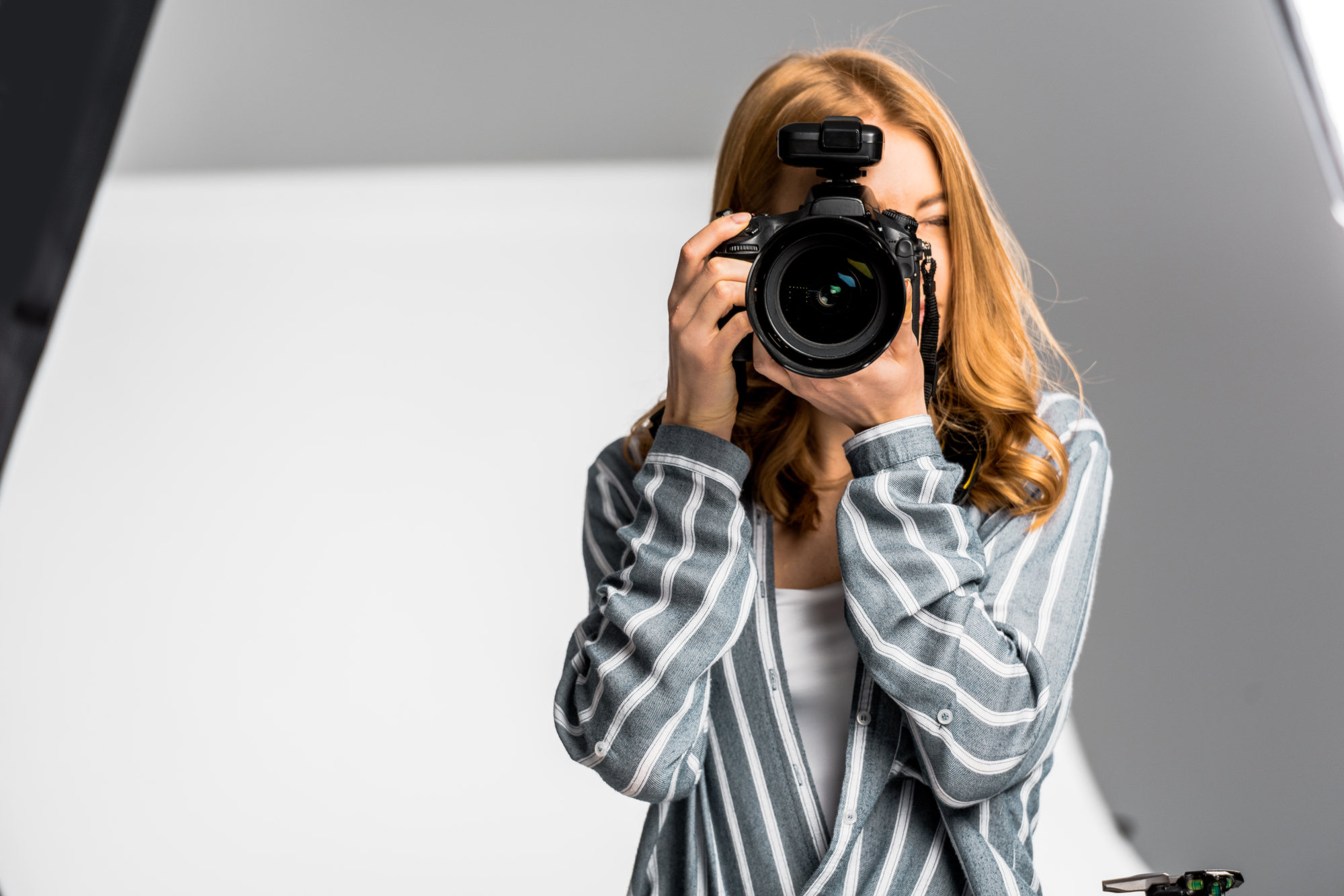 A person with long, light brown hair wearing a striped shirt is holding a camera with both hands and taking a photo in a studio setting, possibly illustrating how you can get paid to take pictures.