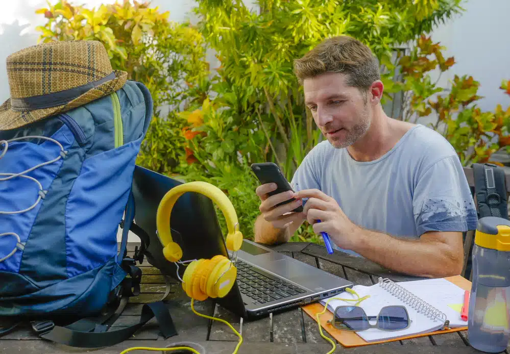 A man sitting at a table outdoors, using his smartphone while surrounded by a laptop, notebook, water bottle, headphones, and backpack. He seems to be exploring options to get paid to move overseas.