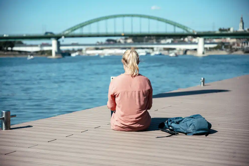 A person with blonde hair sits on a wooden dock by a river, facing away. There is a backpack beside them and a green arch bridge in the background. They seem lost in thought, perhaps dreaming of an adventure where they get paid to move overseas.