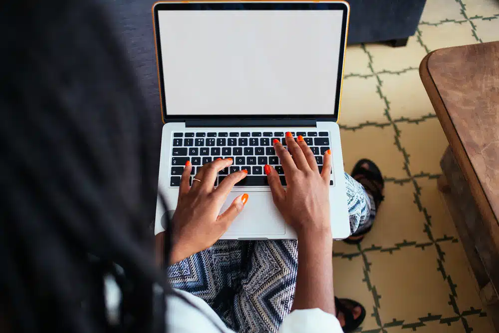 A person with orange painted nails types on a laptop, perhaps researching how to get paid to move overseas. The floor has a geometric pattern and a wooden table is nearby.
