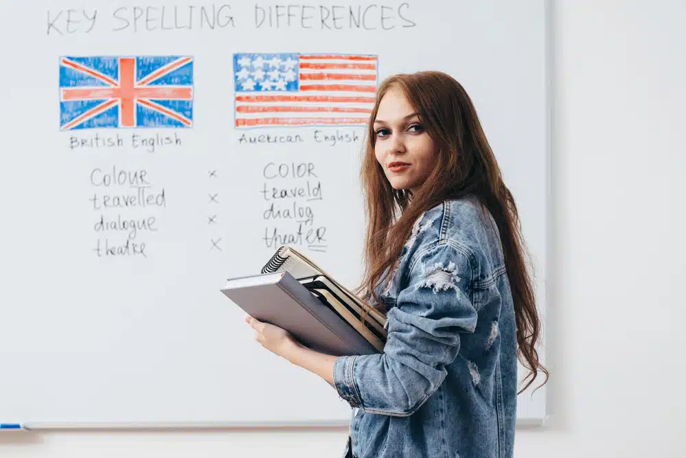 A woman holds books and stands in front of a whiteboard displaying British and American English spelling differences, sharing tips on how to get paid to move overseas.
