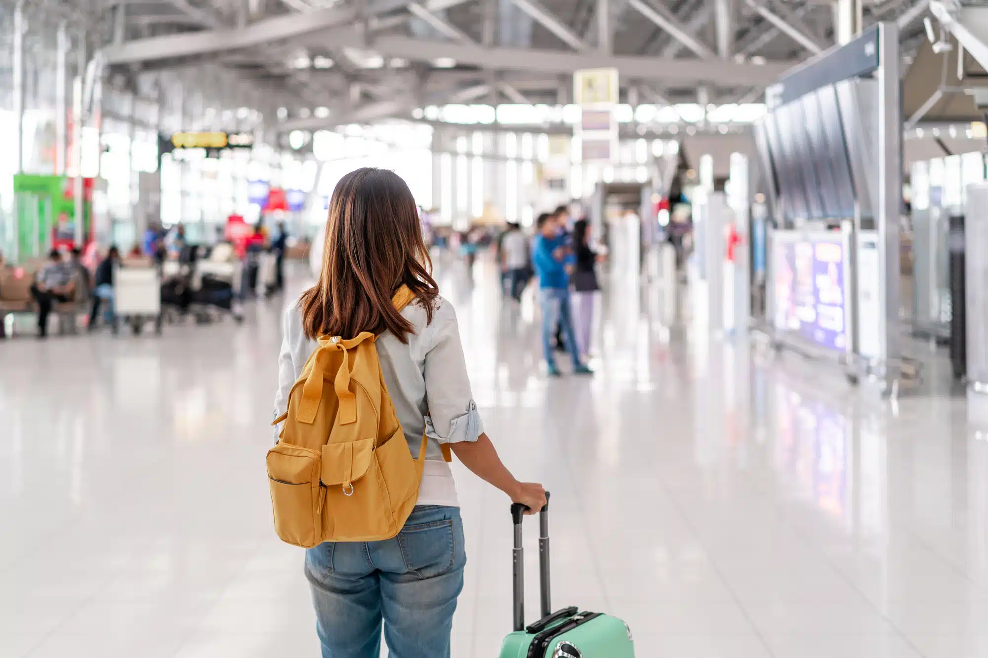 A person with a yellow backpack and a suitcase stands in an airport terminal, facing away, perhaps contemplating how one could get paid to move overseas, while other people and airport signage are visible in the background.